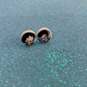Può includere: Un paio di orecchini a bottone rotondi con un'illustrazione a cartoni animati di una persona sorridente con capelli neri e una camicia rossa. Gli orecchini sono su uno sfondo azzurro glitterato.