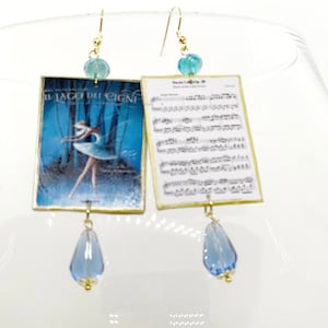 Può includere: Orecchini pendenti color oro. Uno presenta un'immagine di ballerina de "Il Lago dei Cigni" e l'altro spartiti. Ogni orecchino ha una perla blu e un ciondolo blu a forma di goccia.