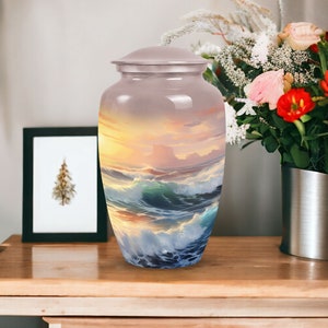 May include: A silver urn with a painted image of a seascape with waves crashing against the shore at sunset. The urn has a lid and a smooth, polished finish.