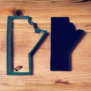 May include: Two cookie cutters in the shape of the Canadian province of Alberta. The cutter on the left is teal blue and the cutter on the right is dark blue.