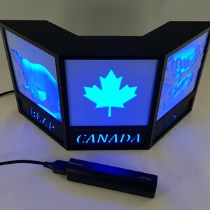 Peut inclure: Lampe LED noire angulaire avec trois panneaux lumineux. Le panneau central affiche une feuille d'érable lumineuse, flanquée de panneaux avec un ours polaire et d'autres images. Le mot "CANADA" est éclairé en dessous. Un câble USB et un adaptateur secteur noirs sont connectés.