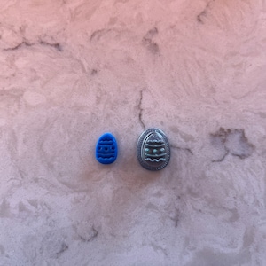 Peut inclure: Deux objets en forme d'œuf de Pâques. L'un est un petit tampon bleu vif avec un motif d'œuf. L'autre est un bouton en métal argenté avec un motif d'œuf. Les deux objets sont sur une surface claire et marbrée.