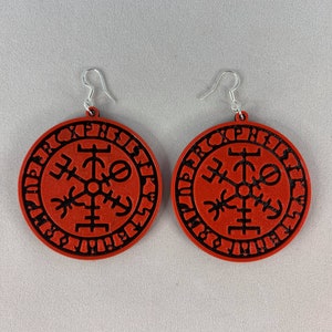 Peut inclure: Une paire de boucles d'oreilles circulaires rouges avec des symboles et des runes noirs. Les boucles d'oreilles sont en matériau texturé et ont des crochets argentés.