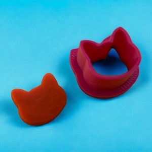 Peut inclure: Un emporte-pièce rouge en forme de chat et un biscuit en forme de chat sur fond bleu. L'emporte-pièce est de forme épaisse et arrondie, et le biscuit est de forme plate et pleine. Les articles sont probablement utilisés pour la pâtisserie.