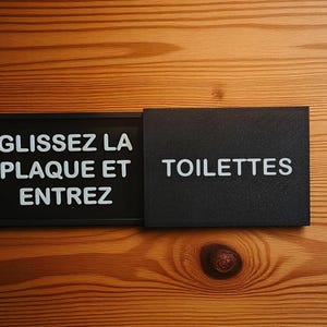 May include: A black rectangular sign with white text that reads "GLISSEZ LA PLAQUE ET ENTREZ" and "TOILETTES". The sign is on a wooden surface with visible wood grain.
