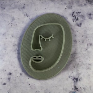 Peut inclure: Un emporte-pièce en forme de visage abstrait vert sauge. Le découpoir présente un design minimaliste d'un œil fermé, d'un nez et de lèvres dans une forme ovale. La surface a un motif texturé. Idéal pour les biscuits.