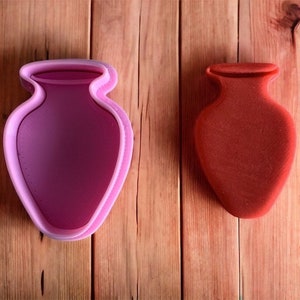 May include: Two vase-shaped cookie cutters on a wooden surface. One is a light pink outline, and the other is a solid red-orange colour. The cutters are designed to create vase-shaped biscuits or decorations.