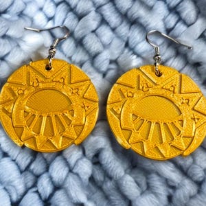 Peut inclure: Une paire de boucles d'oreilles circulaires dorées avec un motif de soleil. Les boucles d'oreilles ont une surface texturée et détaillée et sont suspendues à des crochets d'oreilles argentés. Le motif présente un motif de soleil central avec des lignes rayonnantes et des formes géométriques.