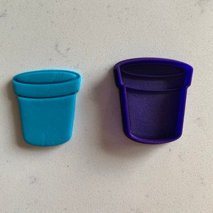 May include: Two plant pot-shaped cookie cutters. One is a solid turquoise blue, and the other is a dark purple outline. The cutters are made of plastic and are designed for baking or crafting. The design is a simple, classic flower pot shape.