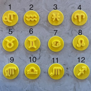 Op de afbeelding: Een verzameling van twaalf gele koekjessnijders met sterrenbeeld symbolen. Elke snijder toont een ander astrologisch teken, waaronder Ram, Stier, Tweelingen, Kreeft, Leeuw, Maagd, Weegschaal, Schorpioen, Boogschutter, Steenbok, Waterman en Vissen. De snijders zijn in een raster gerangschikt.