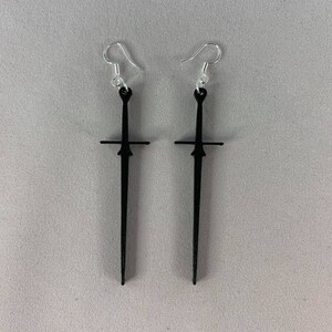 Pode incluir: Um par de brincos pretos em forma de espada com ganchos prateados. Os brincos têm um design de lâmina longo e fino com uma guarda cruzada no topo. Fundo cinza claro.
