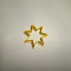 May include: A gold-colored, star-shaped cookie cutter with seven points. The cutter is made of a thin metal and sits on a gray surface. The star shape is a common design for baking and crafting.