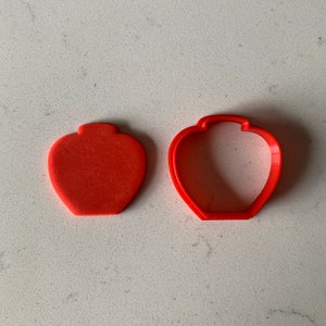 Peut inclure: Deux emporte-pièces orange en forme de vase. L'un est une forme de vase pleine, et l'autre est le contour du vase. Les emporte-pièces sont en plastique et sont sur une surface blanche.