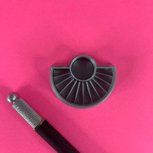 Peut inclure: Une palette grise en forme d'éventail avec des rainures radiales et un puits central circulaire, à côté d'un stylet noir et argenté. Les objets sont posés sur un fond rose vif, créant un contraste saisissant.
