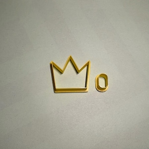 May include: A gold-coloured crown-shaped cookie cutter and a small, rounded rectangle cookie cutter. The crown has five points. The items are on a grey surface.
