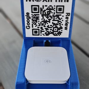 Puede incluir: Un soporte azul impreso en 3D con una parte superior blanca y un código QR para Google Review. El soporte contiene un dispositivo blanco de procesamiento de pagos. El texto "MoxiPrint" está en la parte superior.