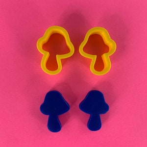 Peut inclure: Deux emporte-pièces en forme de champignon jaunes et deux découpes de champignon bleues sont disposés sur un fond rose. Les emporte-pièces ont un dessus arrondi et une tige. Les découpes sont bleu uni et reproduisent la forme des emporte-pièces.