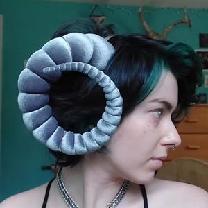 May include: A person wearing a pair of large, gray, velvet ram horns. The horns are curved and textured, with a spiral design. The person is also wearing a silver chain necklace with a lock pendant.