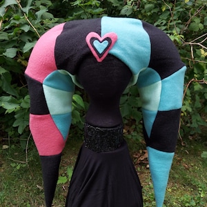 May include: A black, pink, and blue jester hat with a heart-shaped patch on the top. The hat has two long, pointed ends with gold bells at the tips.