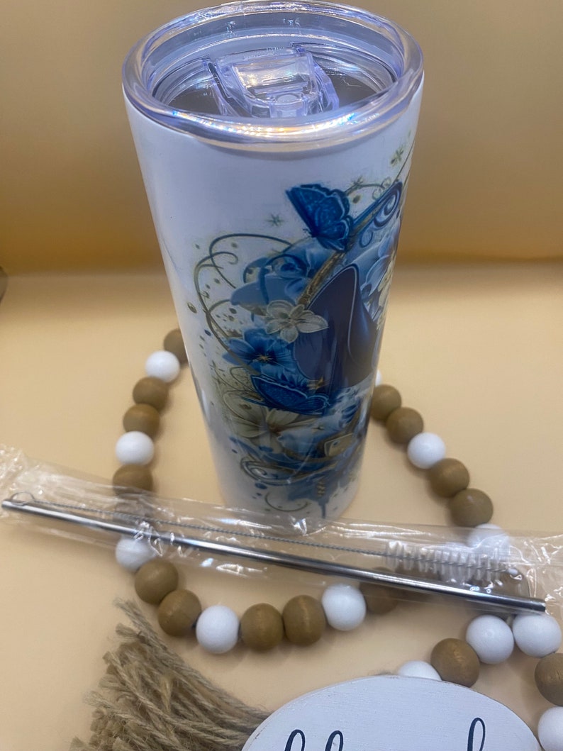 Puede incluir: Un vaso de acero inoxidable blanco con un dise&ntilde;o floral azul y dorado con mariposas. El vaso tiene una tapa transparente y viene con una pajita de acero inoxidable y un cepillo de limpieza.