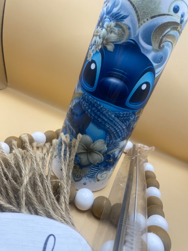 Puede incluir: Un vaso de acero inoxidable azul y blanco con un dise&ntilde;o de personaje de dibujos animados. El personaje es azul con ojos blancos y una bufanda azul. El vaso est&aacute; rodeado de cuentas marrones y blancas.