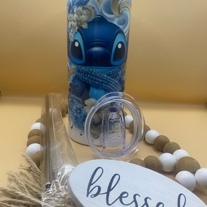 Puede incluir: Un vaso de acero inoxidable azul y blanco con un dise&ntilde;o de personaje de dibujos animados. El vaso tiene una tapa transparente y una pajita. El vaso est&aacute; sobre una mesa con un letrero de madera que dice "blessed".