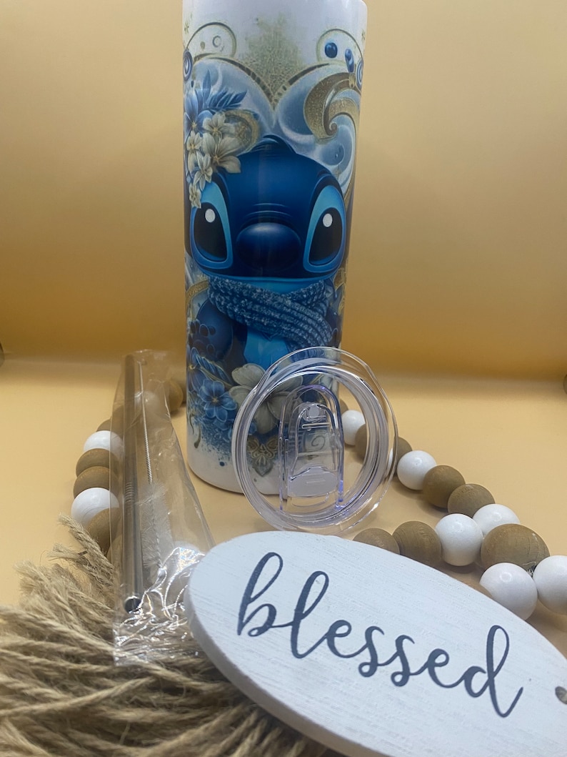 Puede incluir: Un vaso de acero inoxidable azul y blanco con un dise&ntilde;o de personaje de dibujos animados. El vaso tiene una tapa transparente y una pajita. El vaso est&aacute; sobre una mesa con un letrero de madera que dice "blessed".