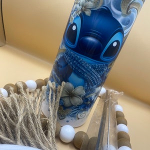 Puede incluir: Un vaso de acero inoxidable azul y blanco con un dise&ntilde;o de personaje de dibujos animados. El personaje es azul con ojos blancos y una bufanda azul. El vaso est&aacute; rodeado de cuentas marrones y blancas.