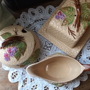 Op de afbeelding: Een set van drie vintage keramische serveerschalen met een druivenrankenontwerp. De schalen zijn beige met bruine ranken, groene bladeren en paarse druiven. De set bevat een roomkan, een suikerpot en een botervloot.