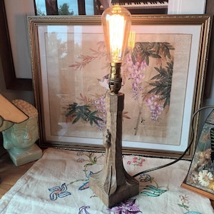 May include: A wooden table lamp with a decorative base and a large Edison-style light bulb. The lamp has a brown finish and a vintage aesthetic. The lamp is plugged into an electrical outlet.