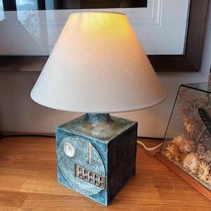 May include: A blue and teal ceramic table lamp with a beige lampshade. The lamp has a square base with a circular design and a rectangular brass-colored detail. The lampshade is cone-shaped. The lamp is on a wooden surface.