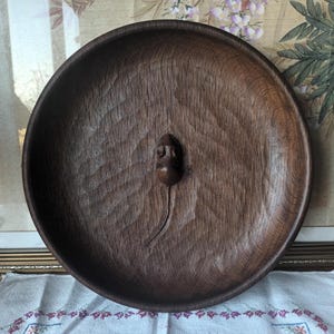 May include: A round, dark brown wooden tray with a carved mouse design in the center. The tray has a textured surface and a raised edge. The mouse has a detailed head and a long, curved tail. The tray is likely handmade.