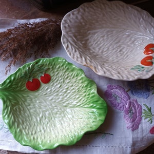 Puede incluir: Dos platos de servir de cerámica con forma de hojas de lechuga. Uno es verde con tomates rojos y el otro es blanco con tomates rojos.