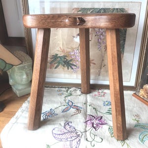 May include: A small, handcrafted wooden stool with three angled legs. The stool is made of light brown wood and features a carved frog detail on the seat. The stool is approximately 12 inches tall.