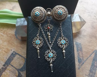 May include: An antique gold-coloured brooch with turquoise and red detailing. The brooch has two circular filigree elements and dangling chains with floral pendants. A distinctive piece of jewellery.