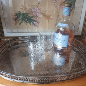 Puede incluir: Una bandeja ovalada plateada con detalles ornamentales contiene dos vasos de cristal y una botella de Dalwhinnie Winter's Gold. La botella tiene una etiqueta azul y dorada. El fondo incluye obras de arte enmarcadas.