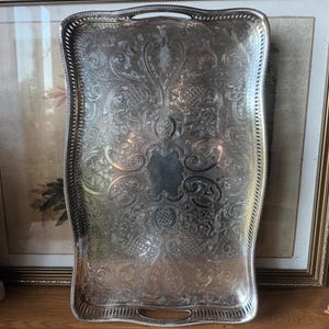 Può includere: Un vassoio da portata rettangolare color argento con un intricato motivo floreale e a volute in rilievo su tutta la superficie. Il vassoio presenta un bordo rialzato traforato e due manici integrati sui lati superiore e inferiore.