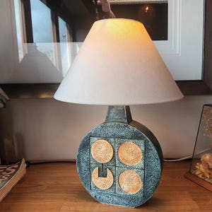 May include: A ceramic table lamp with a white lampshade. The base is a textured blue-gray with four tan circles and a tan square. The lamp is on a wooden surface.