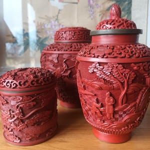 Pode incluir: Três potes decorativos vermelhos, intrincadamente esculpidos com tampas. Os potes apresentam esculturas em relevo detalhadas de paisagens, figuras e padrões florais. O pote maior tem formato de vaso, os outros são cilíndricos.
