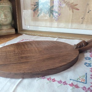 May include: Oval wooden serving board with a handle. The board is a rich brown color and features a carved squirrel on the handle. The board is placed on a white cloth with floral embroidery. A decorative item for the kitchen.