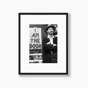 May include: A black and white photograph of a man wearing a hat and a jacket standing in front of a sign that reads "I AM THE DOOR".