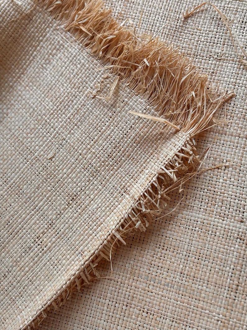May include: Close-up of a woven natural fiber textile with a fringed edge. The fabric has a tight, square weave in a light beige color. The fringed edge is made of loose, straw-like fibers.
