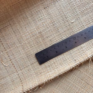 May include: Close-up of a woven natural fiber material with a ruler. The ruler, marked in inches, is positioned diagonally across the textured surface. The material has a light tan color and a visible woven pattern.