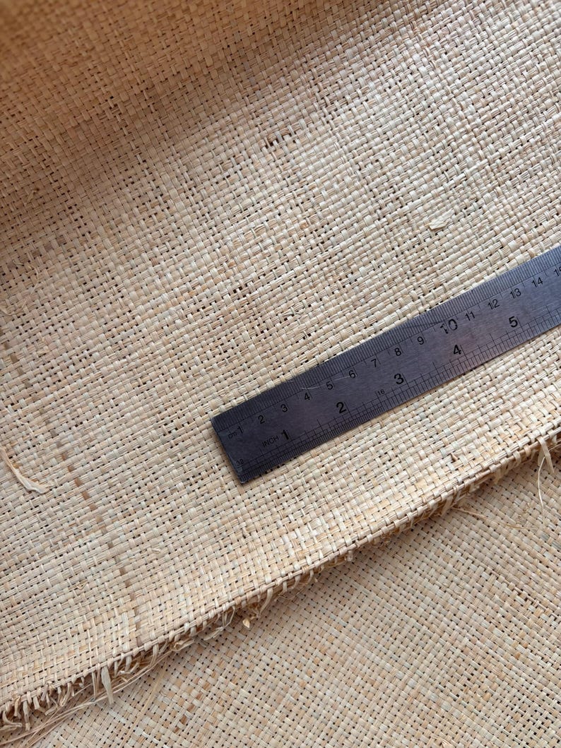 May include: Close-up of a woven natural fiber material with a ruler. The ruler, marked in inches, is positioned diagonally across the textured surface. The material has a light tan color and a visible woven pattern.