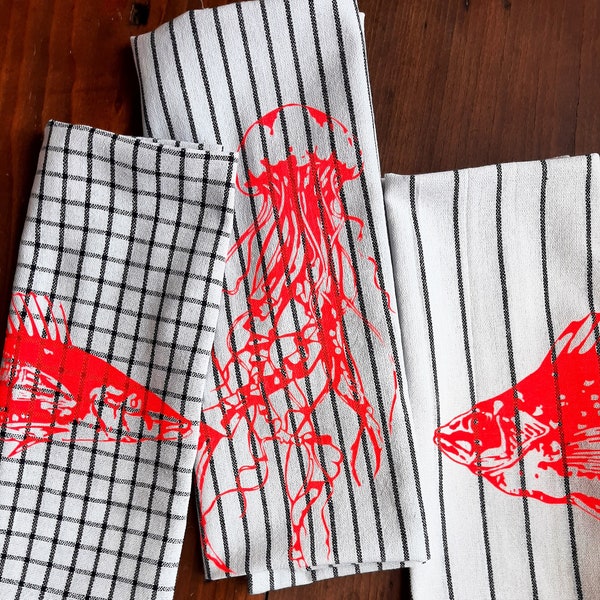 Puede incluir: Conjunto de tres toallas de cocina con temática náutica. Las toallas presentan un patrón de rayas blancas y negras con diseños gráficos rojos de una medusa, un pez y un caballito de mar.