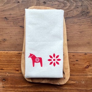 Könnte beinhalten: Gefaltetes weißes Küchentuch mit rotem Dala-Pferd und Blumenmuster. Das Tuch besteht aus einem hellen Stoff und liegt auf einem Holzschneidebrett. Der Hintergrund ist eine Holzoberfläche.