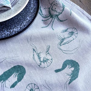 Op de afbeelding: Een lichtpaarse linnen tafelkleed met een teal krab- en garnalenontwerp. Het tafelkleed wordt gecombineerd met een blauwe, gespikkelde plaat en een bijpassende servet. Het ontwerp heeft een nautisch thema.