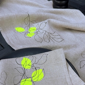 Può includere: Runner da tavolo in lino beige e set di tovaglioli coordinati. Il runner e il tovagliolo presentano un disegno a tratto nero di un motivo floreale con vivaci accenti giallo neon. Gli articoli sono esposti su una superficie nera.