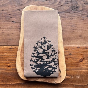 May include: A brown linen tea towel with a black silhouette of a pine cone printed on it. The tea towel is folded and placed on a wooden surface.