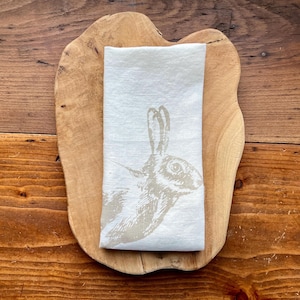 Può includere: Un tovagliolo di lino bianco con un'illustrazione di coniglio marrone stampata su di esso. Il tovagliolo è piegato e posizionato su un vassoio di servizio in legno.